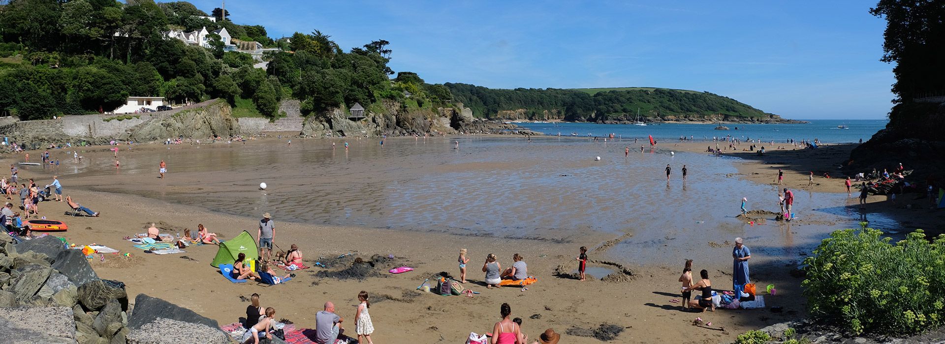 People on a sandy beach. Lush green trees line the coast. Clear blue sky.