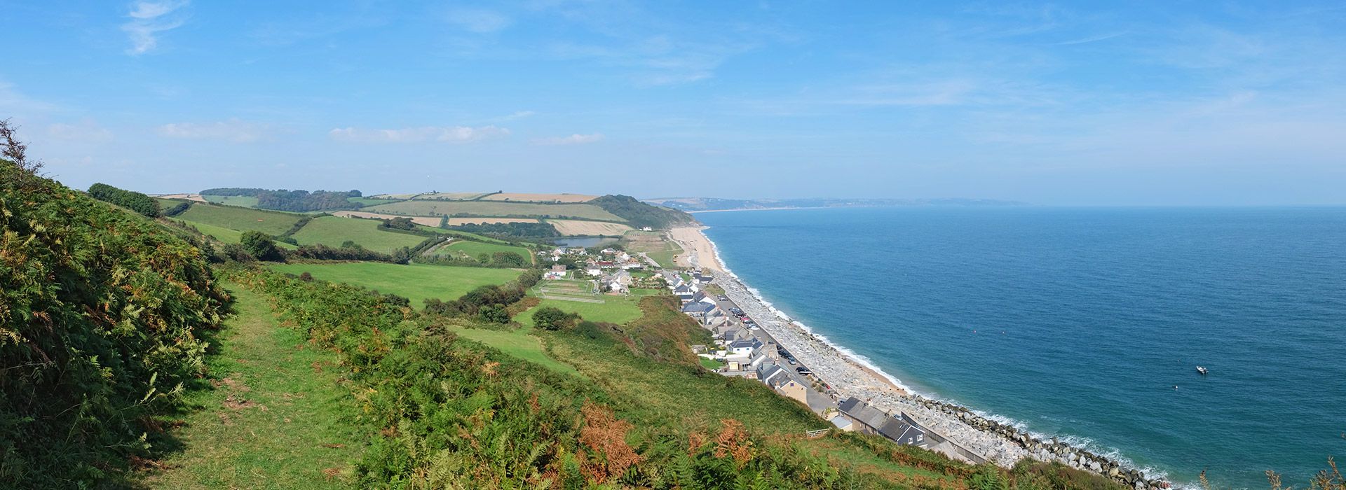 Coastal view with green hills, a pebble beach, and the blue ocean under a clear sky.