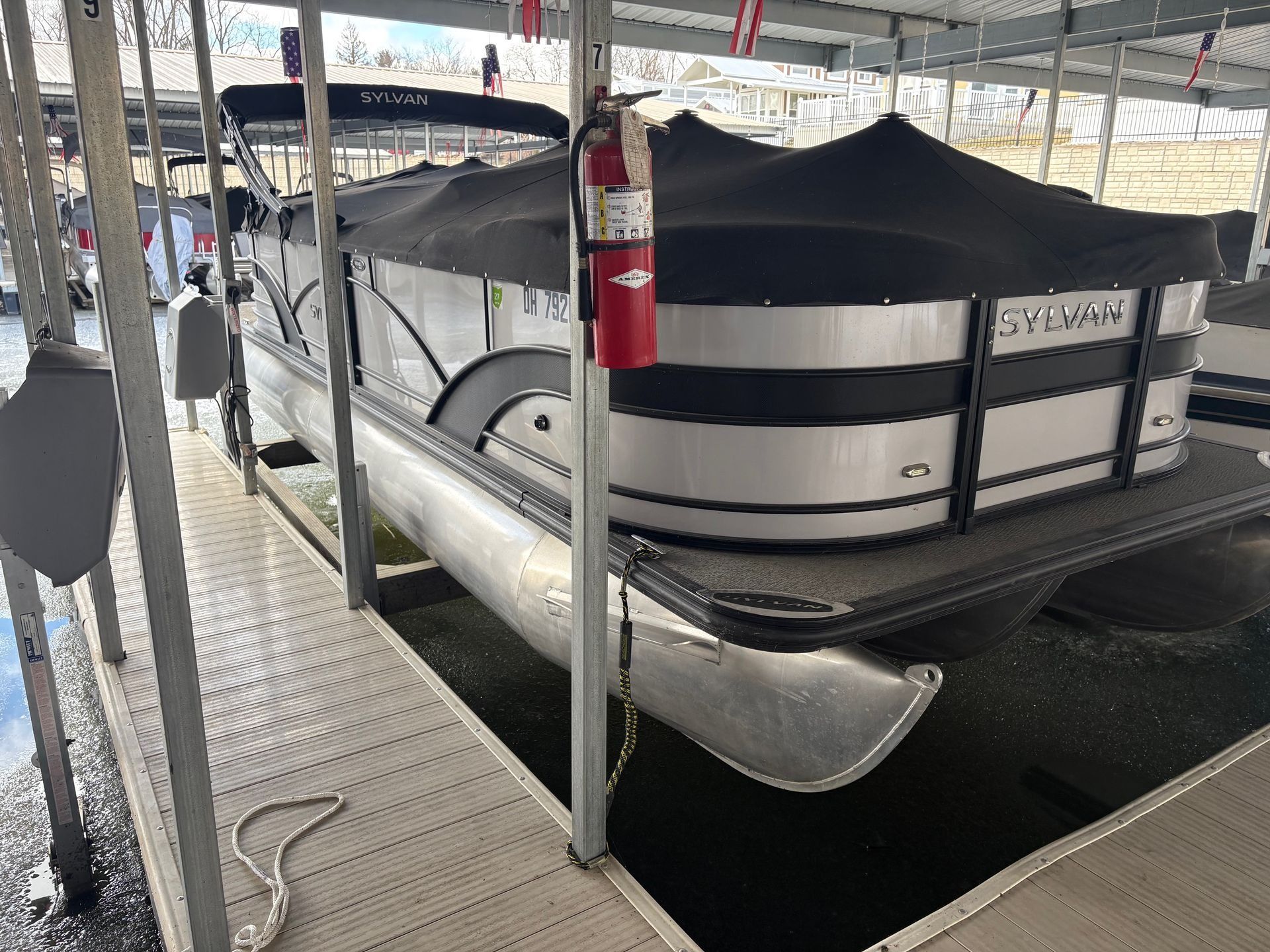 Pontoon boat covered by a black canopy on a trailer under a metal shelter, with a red fire extinguisher nearby.