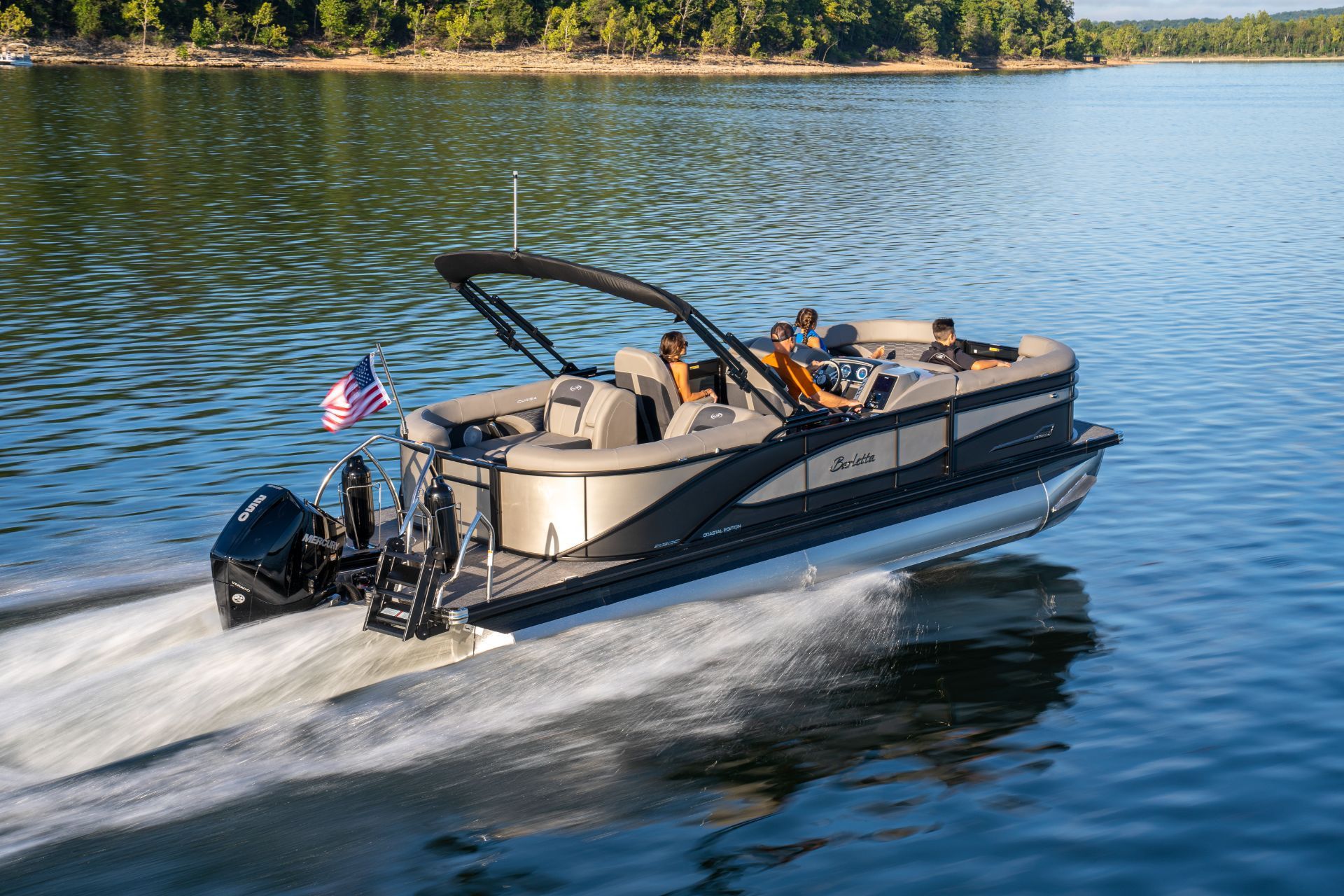 Pontoon boat speeding across a lake, leaving a wake near a wooded shoreline.