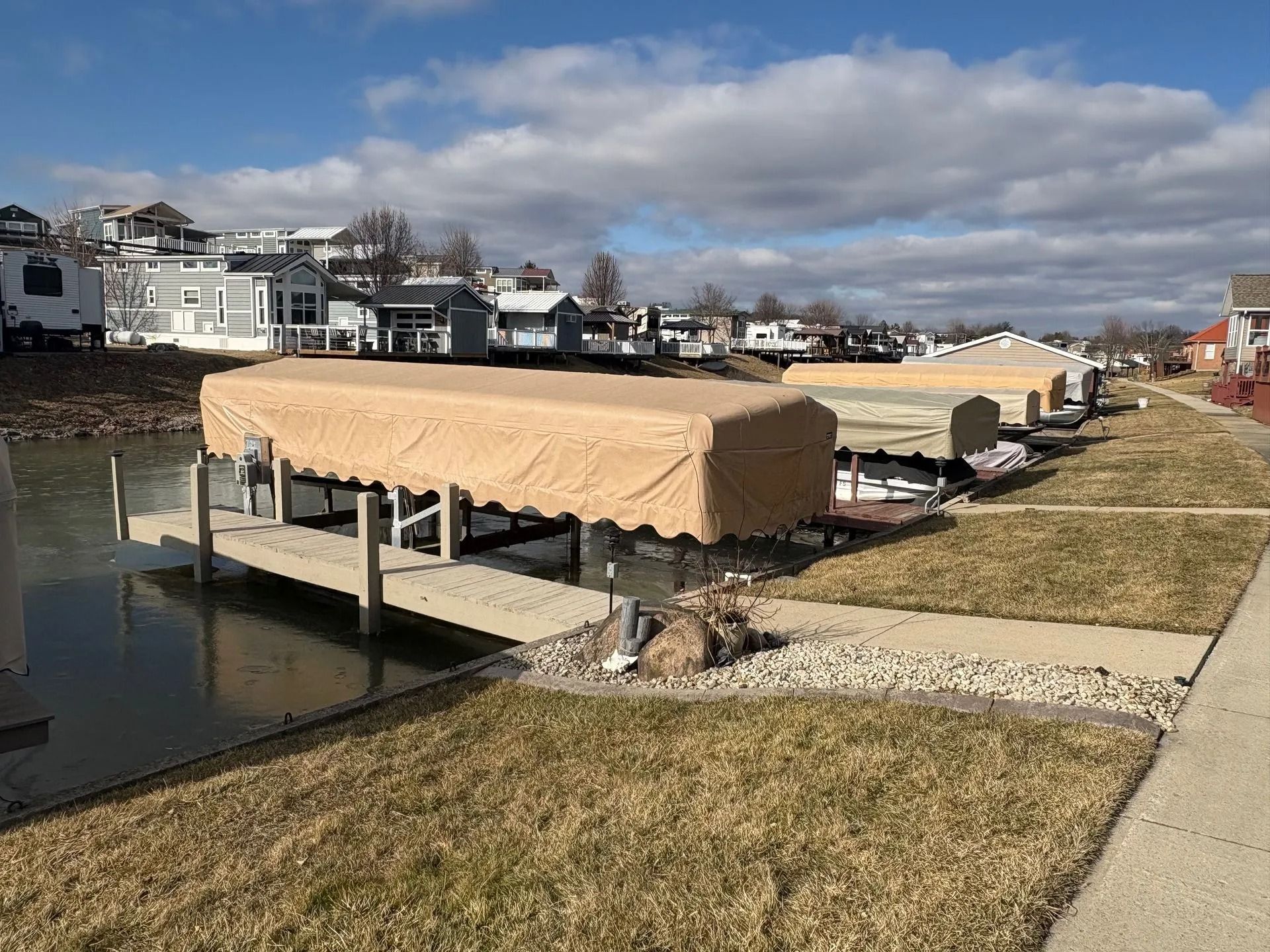 Covered boat lift on a calm canal beside a grassy path and waterfront houses under cloudy skies