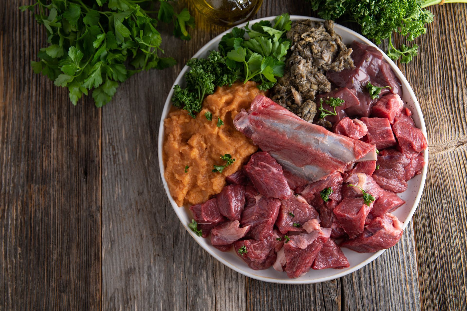 Teller mit rohem Hundefutter: Fleisch, Innereien, Gemüse und Grünzeug auf einem Holztisch.
