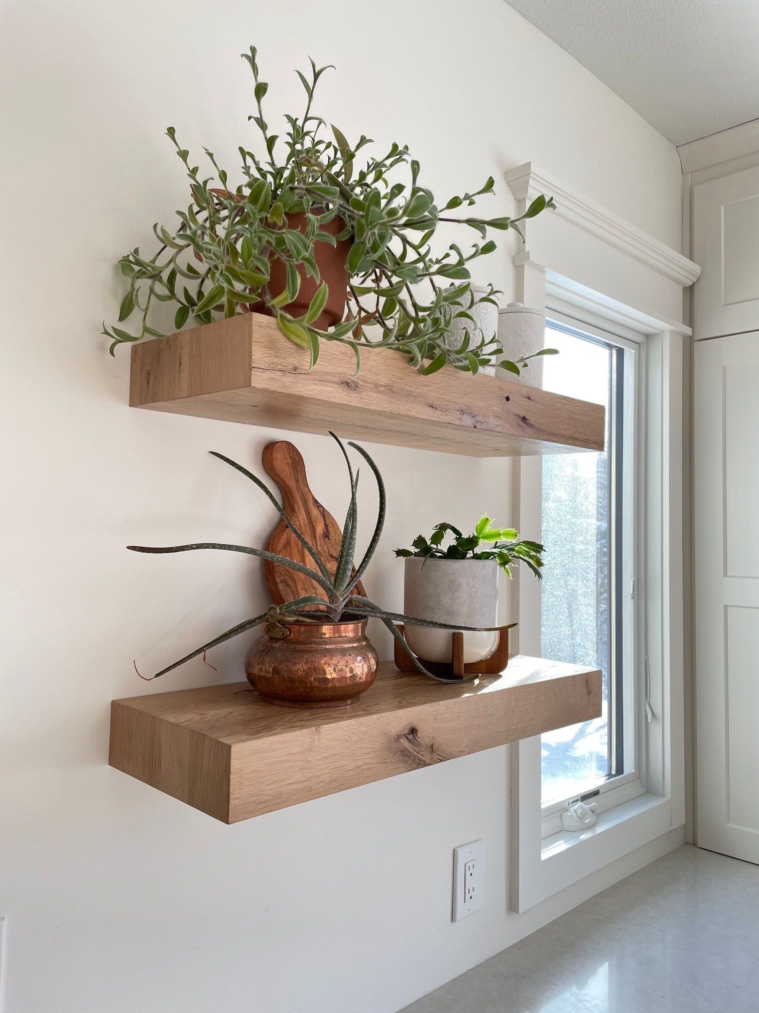 Two wooden shelves with potted plants on them are hanging on a white wall next to a window.