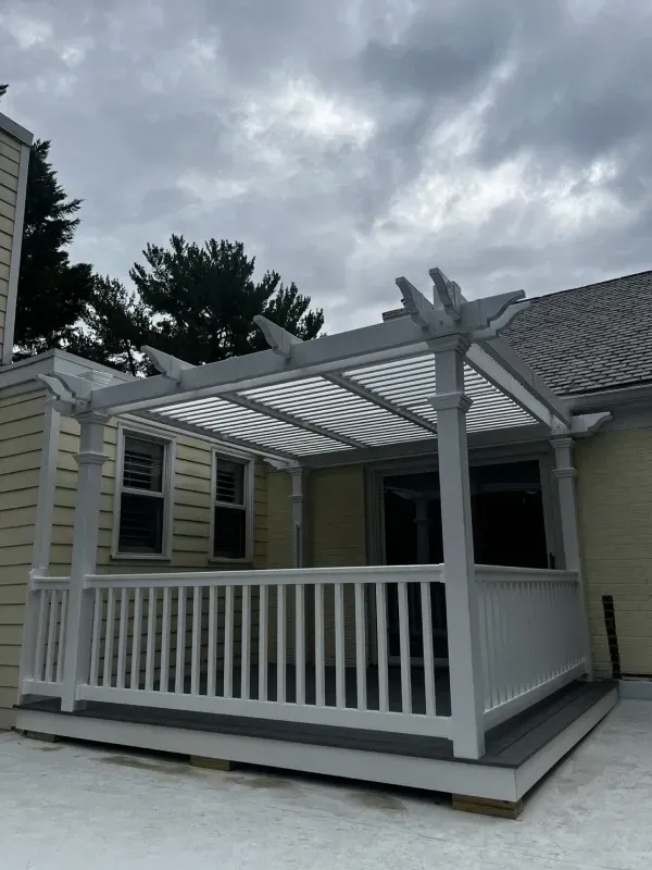 White pergola over a deck attached to a yellow house, under a cloudy sky.