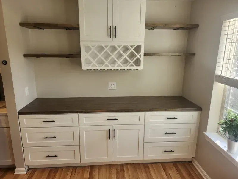 Built-in kitchen storage with white cabinets, dark countertop, open shelving, wine rack, and window.