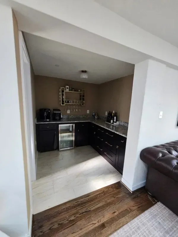 Dark wood cabinets and a mini-fridge in a small kitchen. Tan walls, stone countertop, and wood floor.