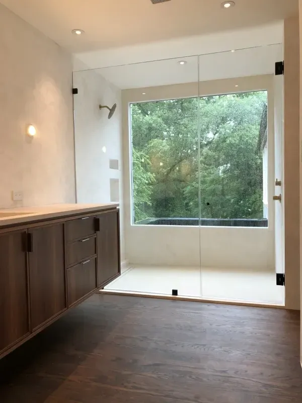Modern bathroom with glass shower overlooking greenery; brown vanity; dark wood floor.