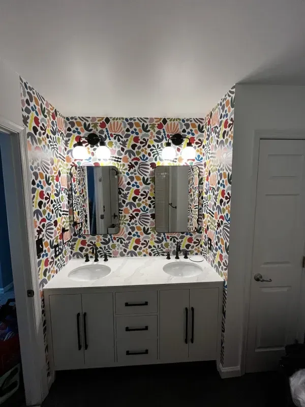 Bathroom with double vanity, two mirrors, and patterned wallpaper. White cabinet with black hardware.