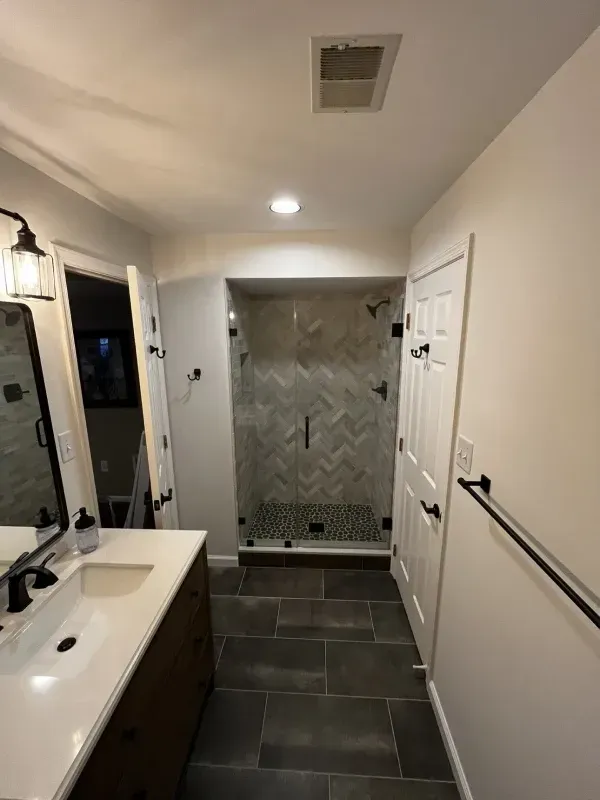 Modern bathroom with walk-in shower. Gray and white herringbone tile, dark wood vanity, black fixtures, and white walls.