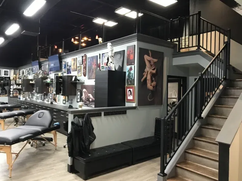 Tattoo studio interior with tattoo stations, art on walls, and a staircase leading to an upper level.