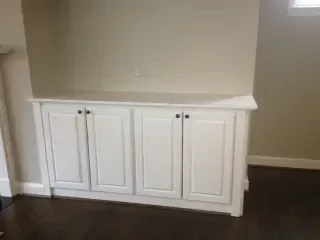 White cabinet with four doors built into a wall, set against neutral beige paint; dark wood floor.