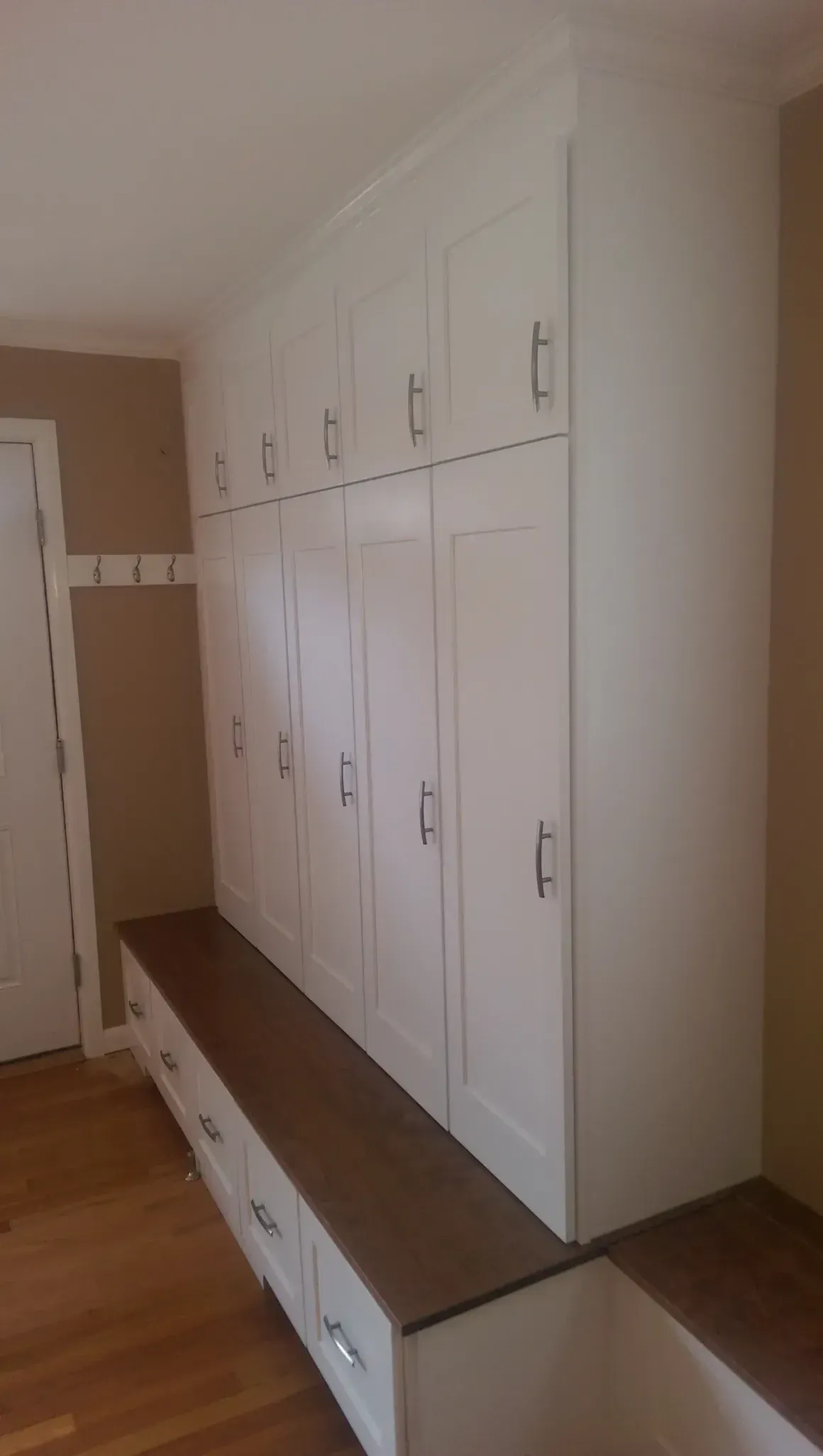White built-in storage unit with cabinets, hooks, and bench; brown countertop and light wood floor.