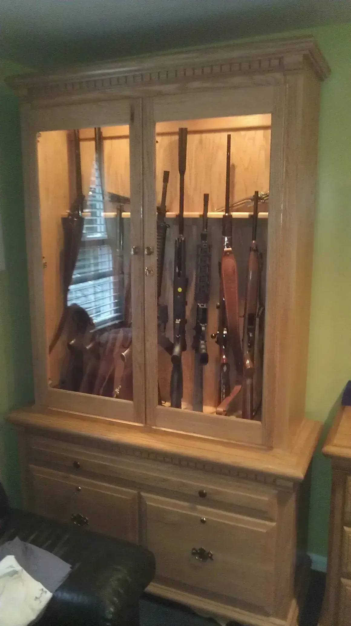 Wooden gun cabinet with glass doors, illuminated interior, holding several rifles.