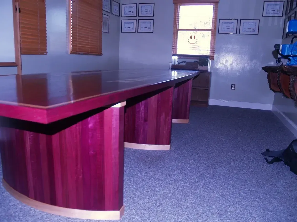 Conference room with large, purple wooden table. Brown blinds, window, blue carpet, awards on wall.