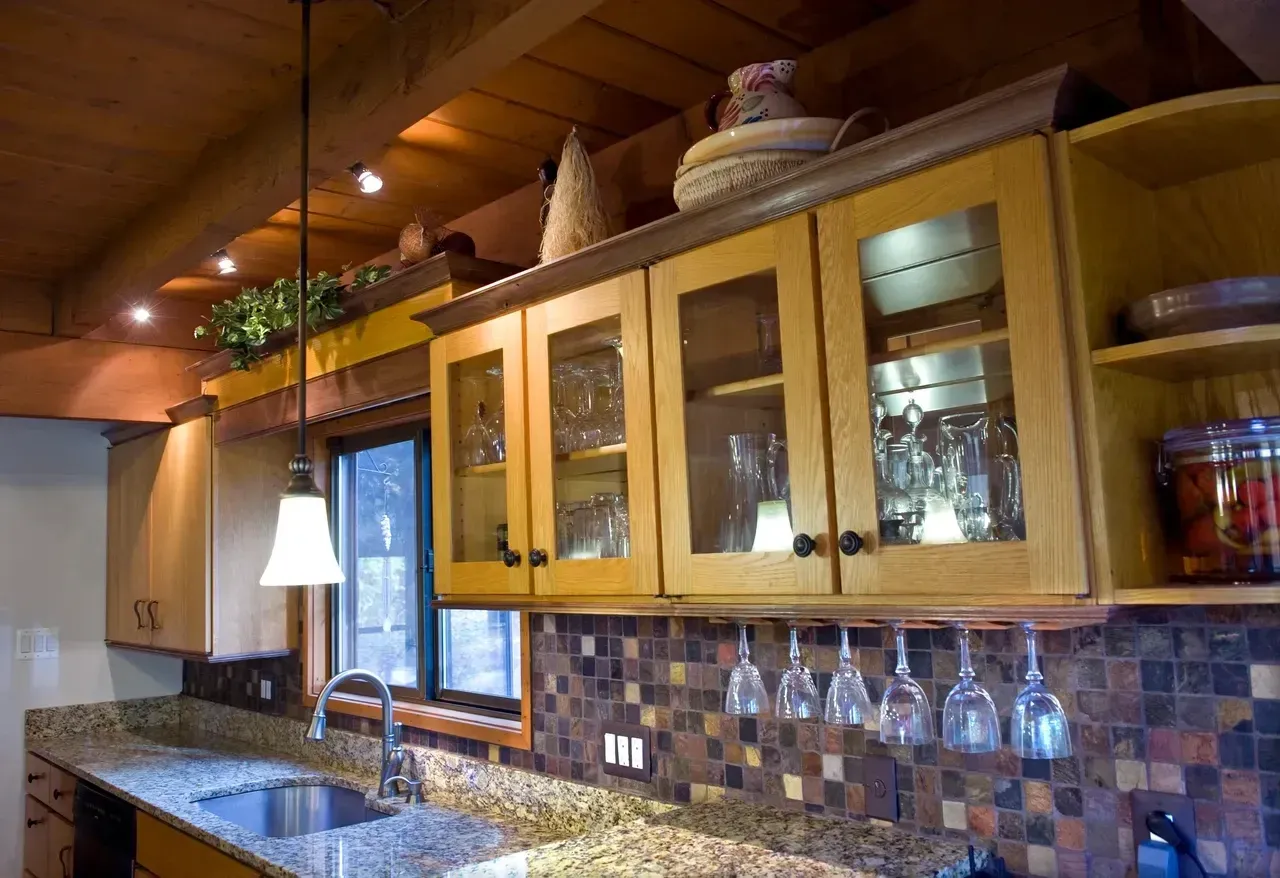 Kitchen with light wood cabinets, granite countertop, mosaic backsplash, and hanging wine glasses.