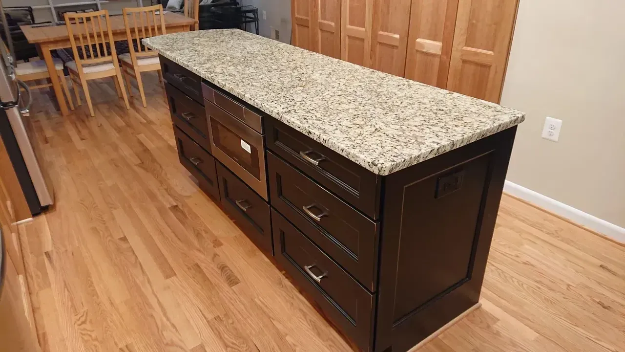 Kitchen island with granite countertop, dark cabinets, built-in microwave, wood floor.