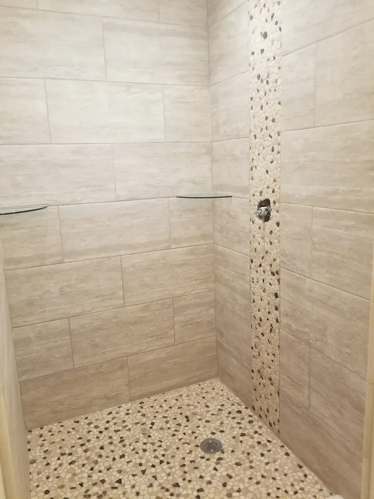 Tiled shower with pebble floor, stone accent strip, and glass shelves. Beige and brown tones.