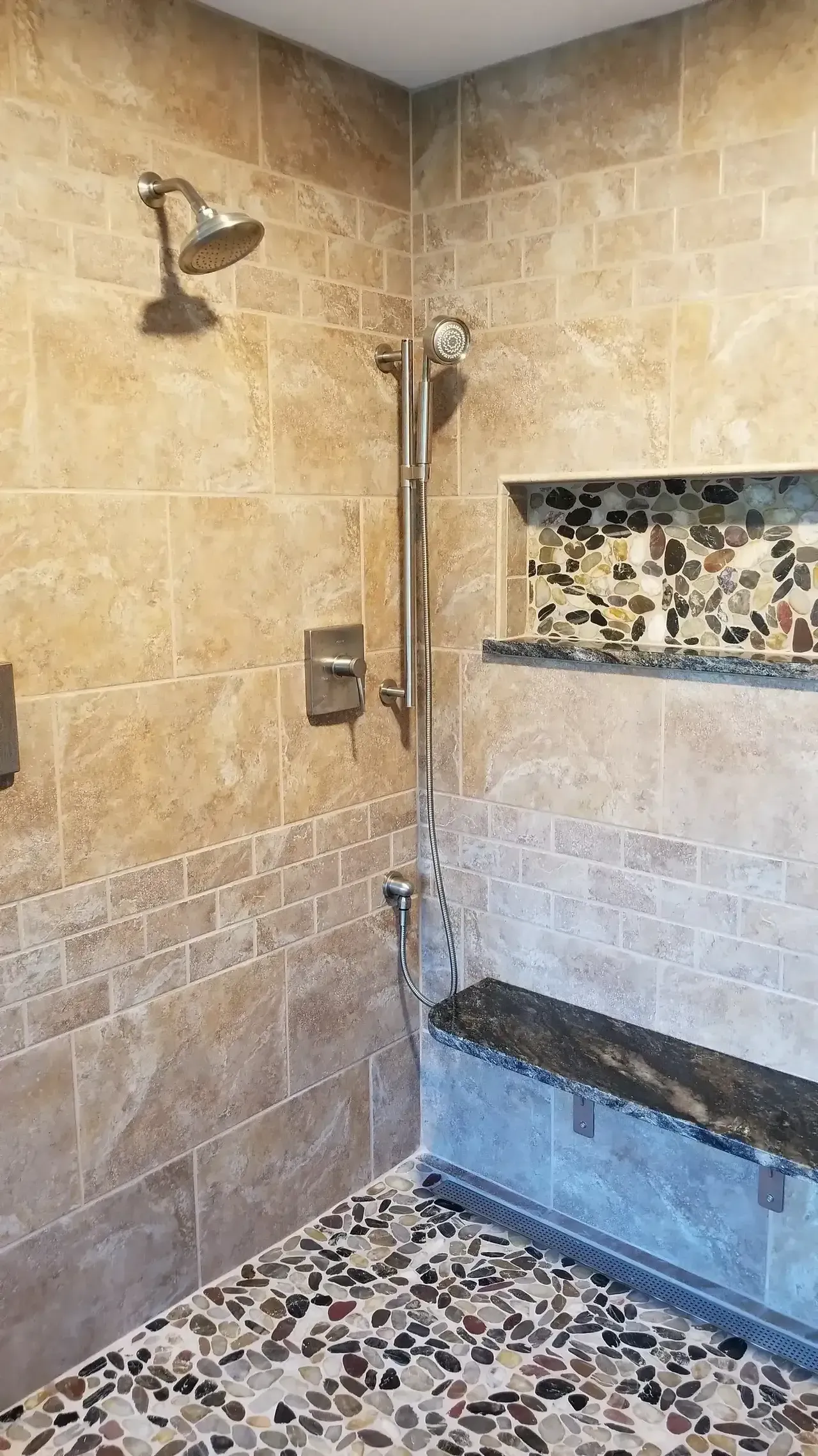 Shower with beige tile walls, pebble floor, built-in bench, and showerheads.