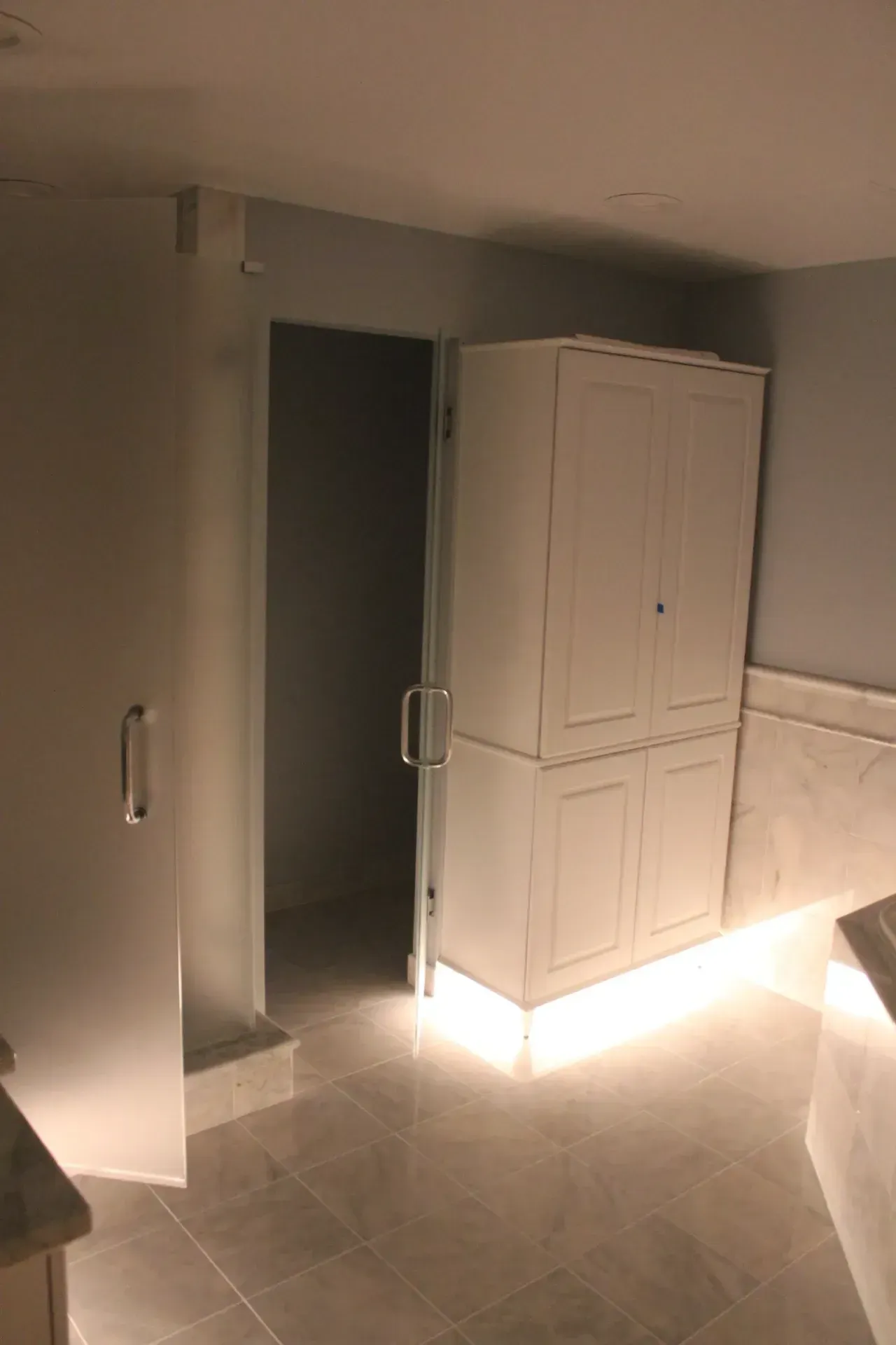 Bathroom with a frosted glass shower door, open door, and white cabinet with under-cabinet lighting.
