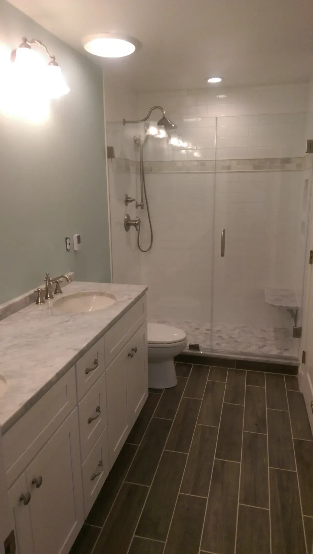 Bathroom with white vanity, large shower, and dark wood-look floor tiles. Light blue wall and bright lighting.
