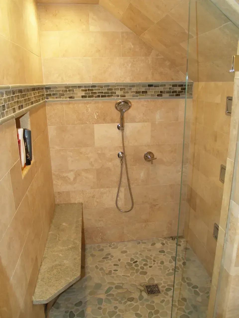 Shower with stone tile walls, pebble floor, glass door, and built-in bench.