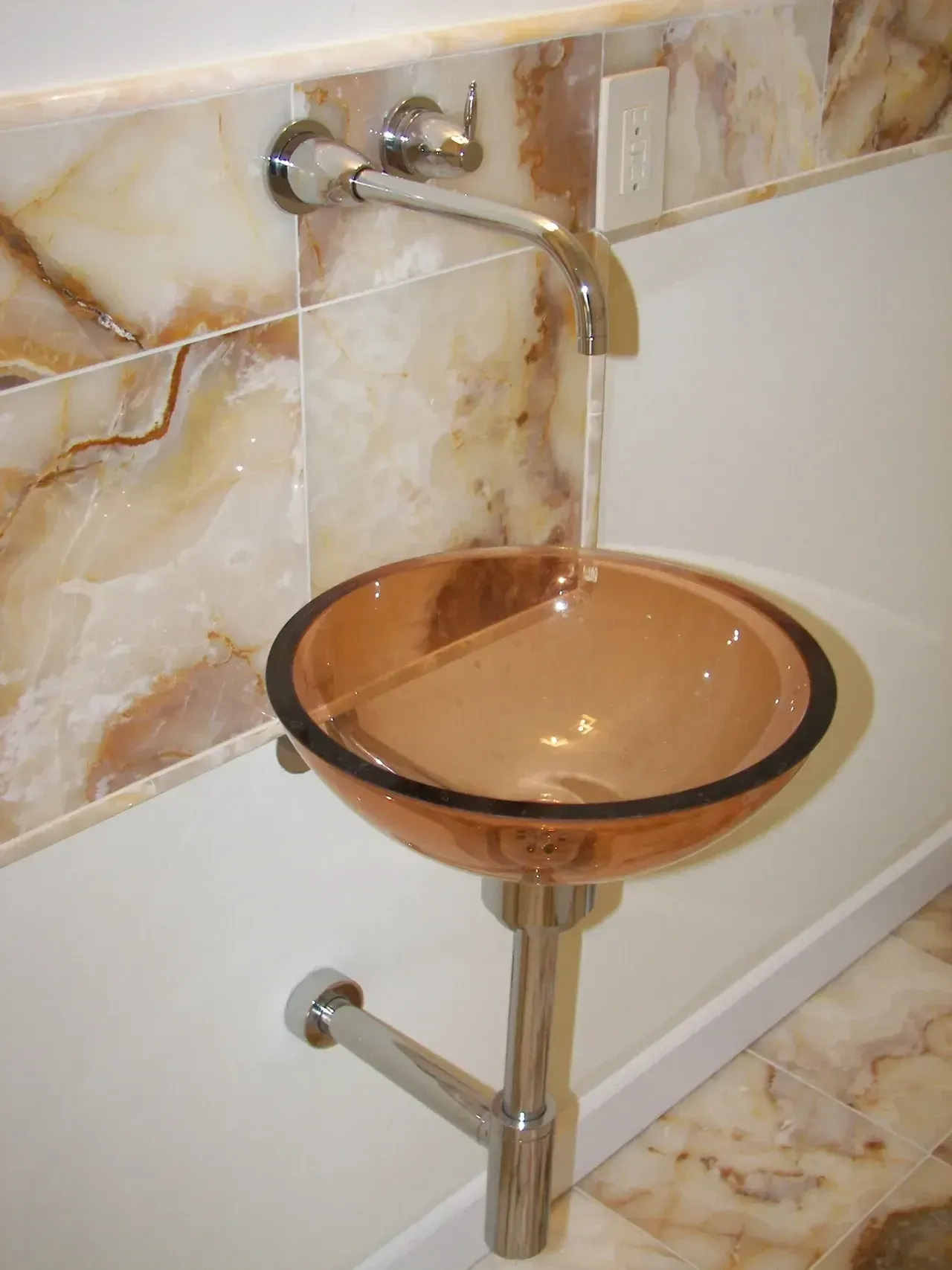 Wall-mounted sink with orange glass bowl and chrome faucet. Tile and flooring are beige with brown veining.