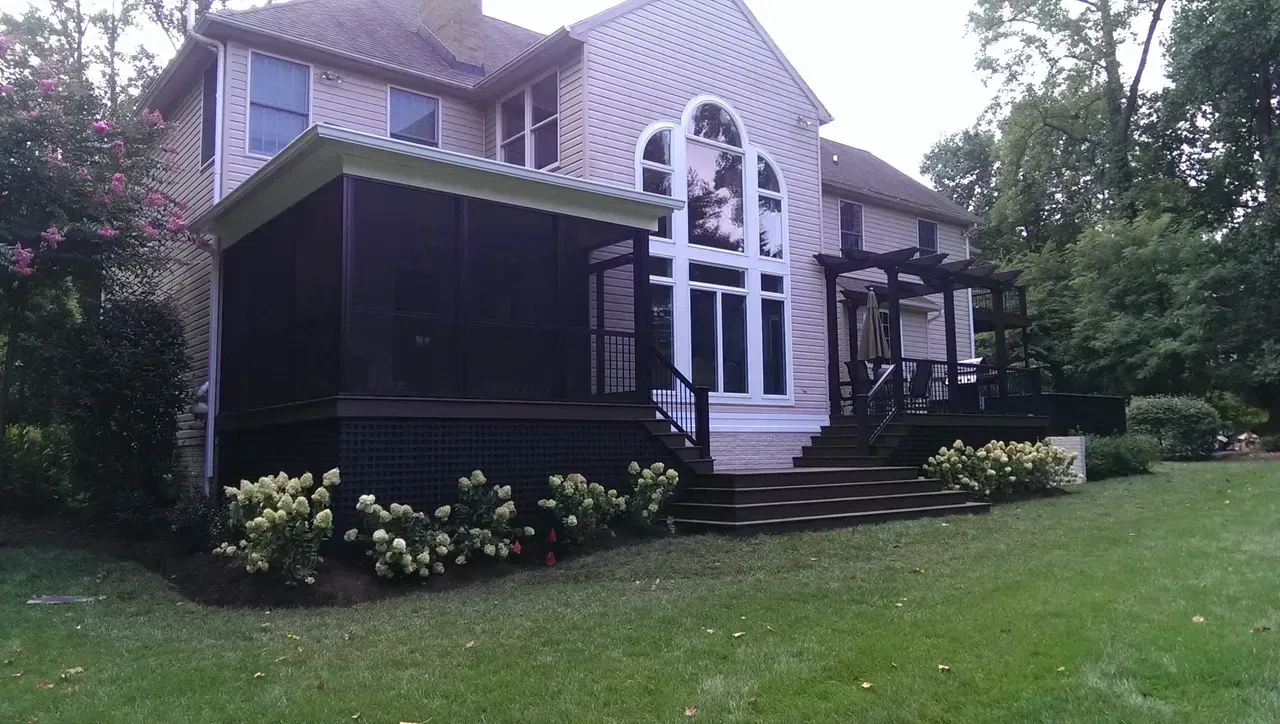 Back of a two-story beige house with a screened-in porch, deck, and pergola, surrounded by greenery and a grassy yard.