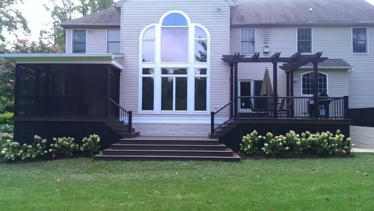 Backyard with a house, steps, decks, a screened porch, and a pergola. Landscaping includes green lawn and white flowers.