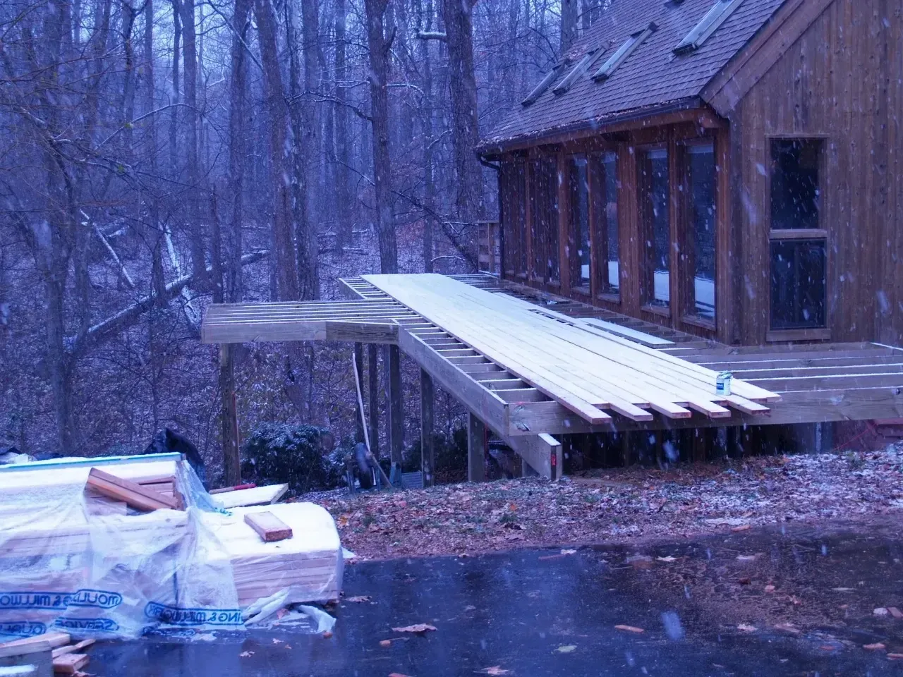 Snow falling on a house with a deck holding building materials; woods in background.