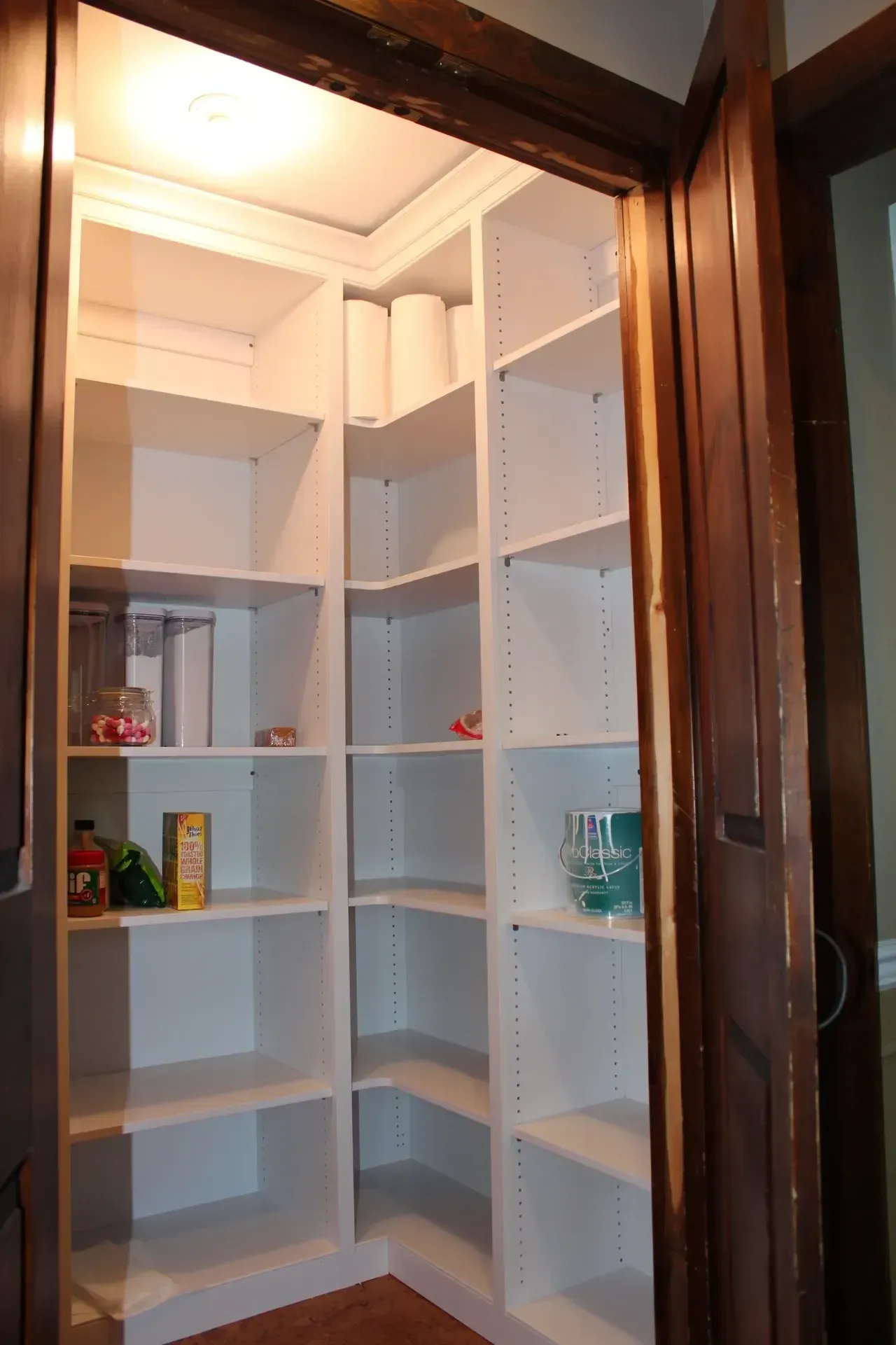 Corner pantry with white shelves, brown door frame, and items like jars and paper towels.