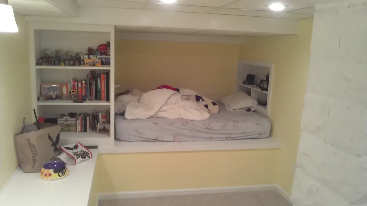Built-in bed nook with shelves and a bed, painted yellow, and white trim. Toys, books, and bedding visible.
