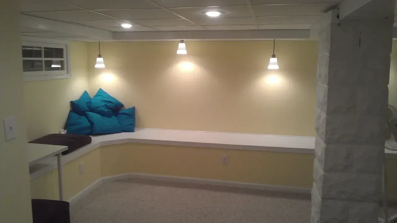 Basement seating area with yellow walls, white bench, blue pillows, pendant lights.