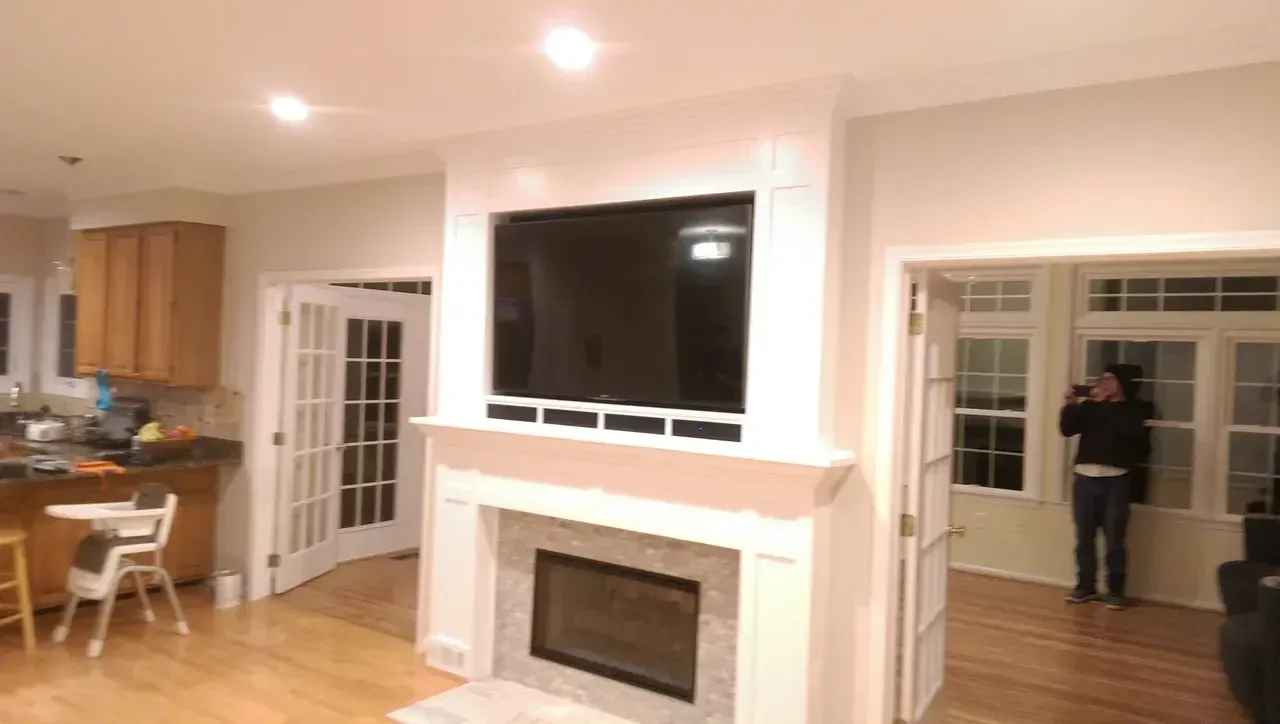 A living room with a TV mounted above a fireplace, and a person in a doorway with windows.