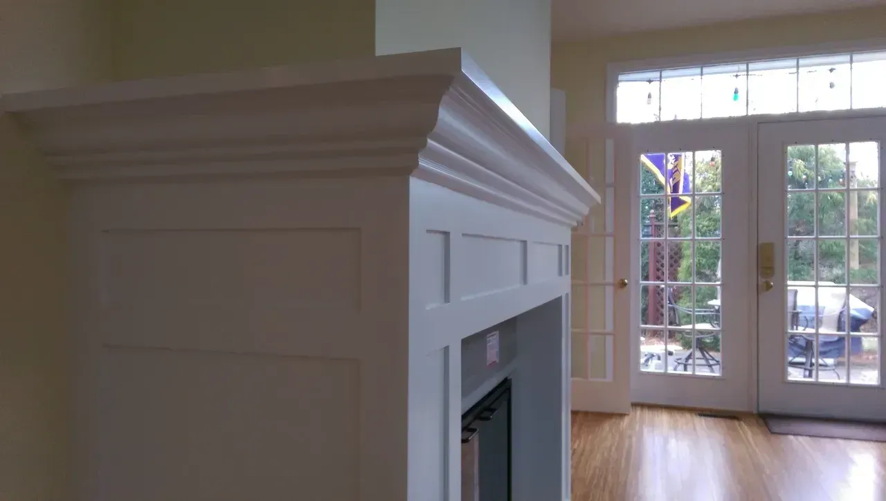 White fireplace mantel with layered molding. Double glass doors in background, hardwood floor.