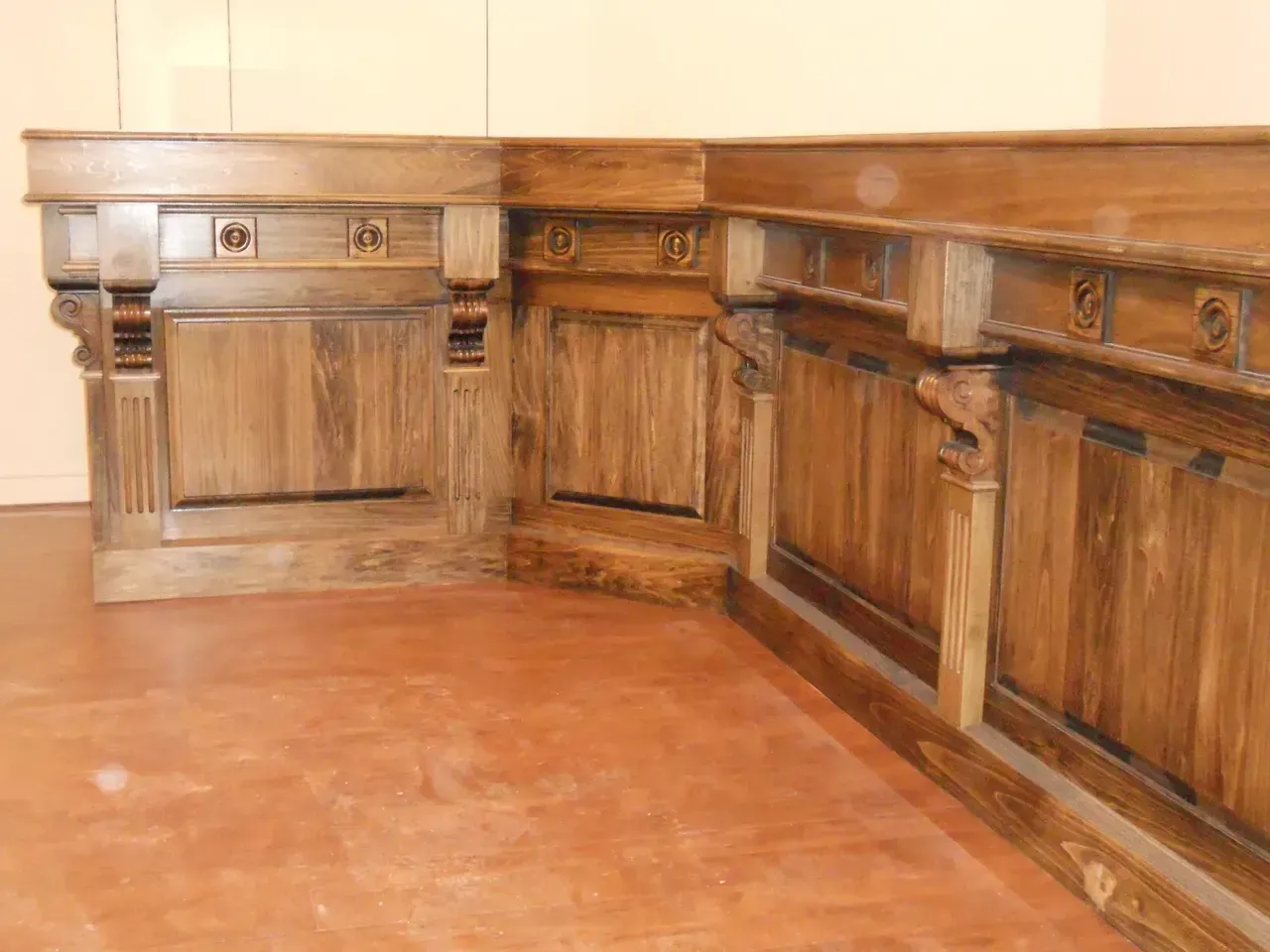 Corner wooden desk, with detailed carvings, resting on a reddish floor.