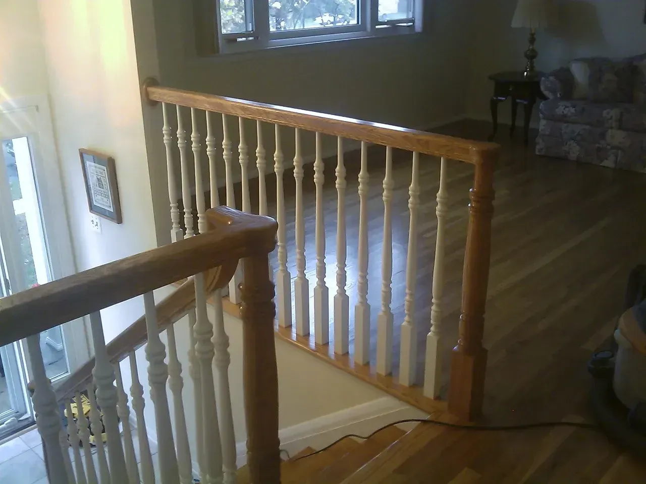 Wooden stair railing with white spindles and tan top rail. Sunlight from window.