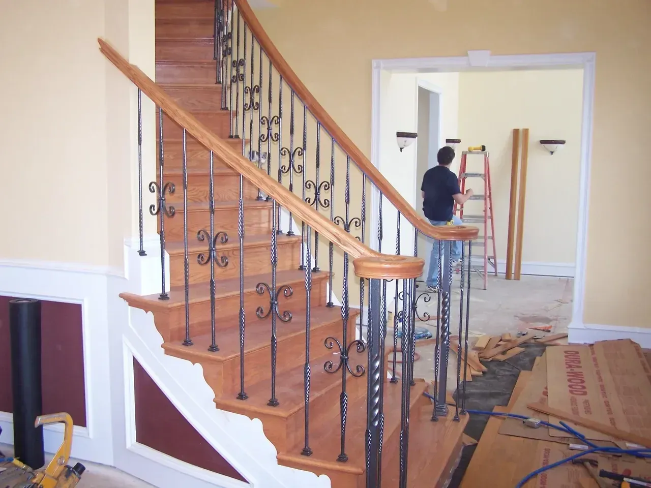 Wooden staircase with black wrought iron balusters, person in background.