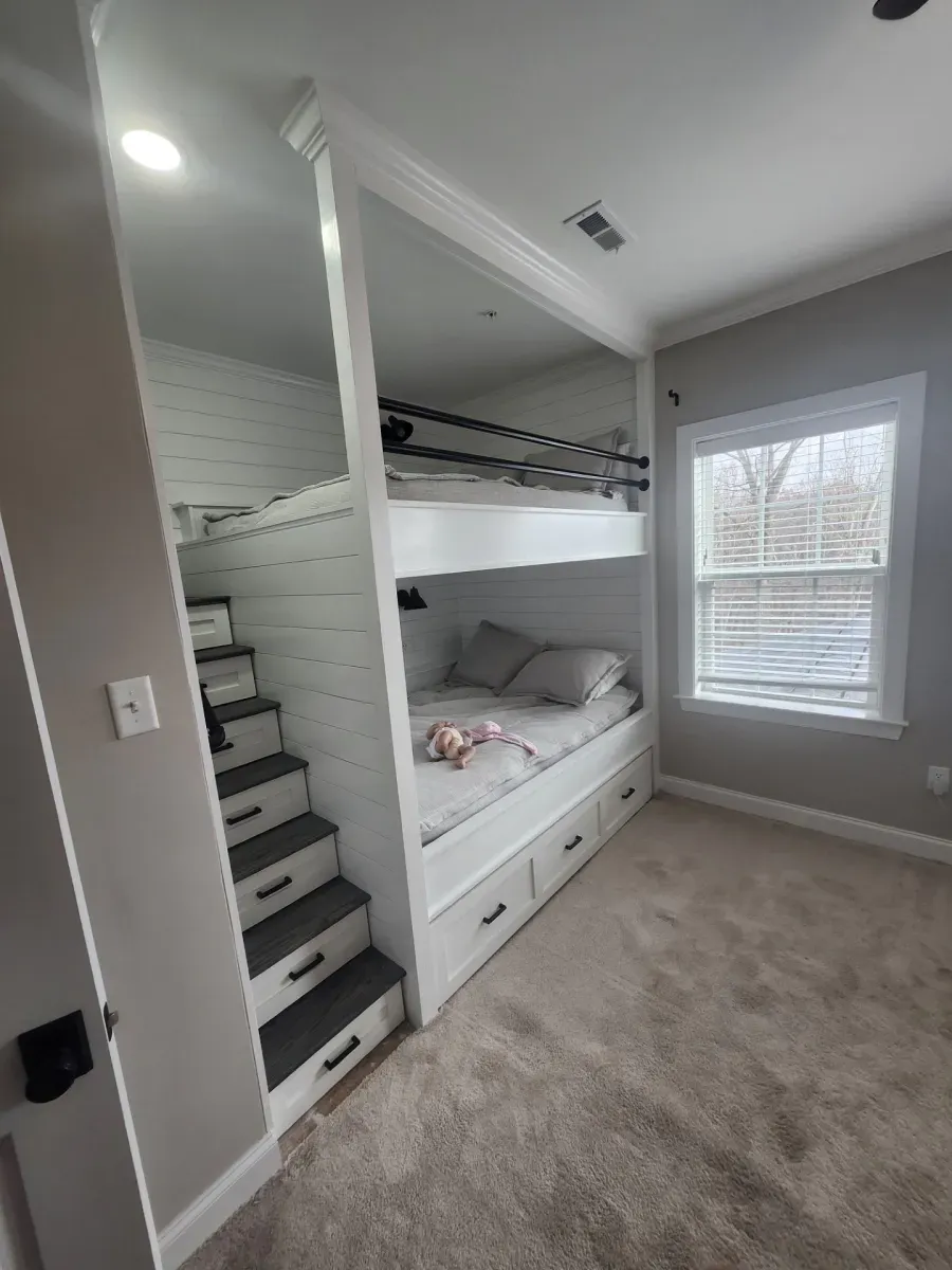 Built-in white bunk beds with drawers and stairs in a bedroom with a window and carpet.