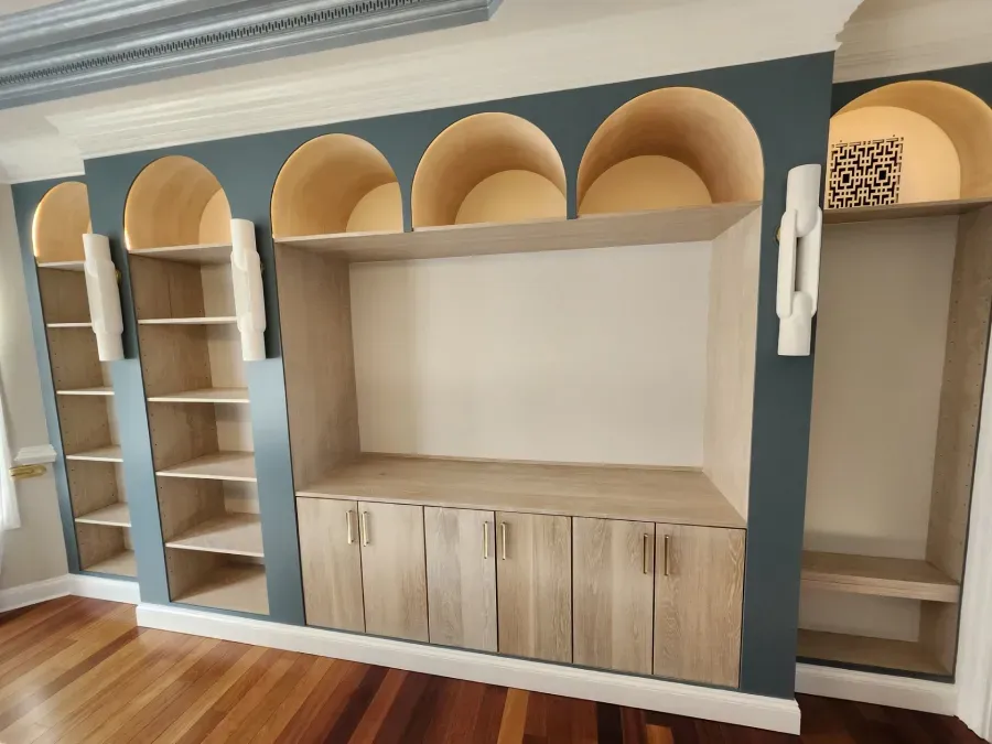 Built-in wooden shelving with arched cutouts and cabinets against a teal wall. The floor is hardwood.