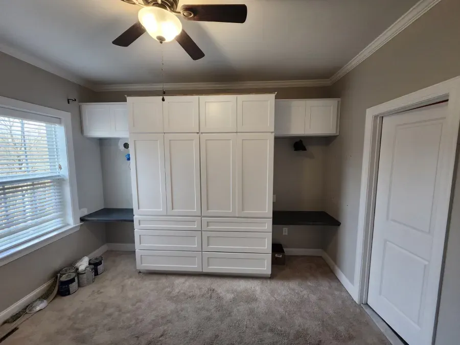 White built-in storage unit with drawers and cabinets, flanked by dark gray desks, near a window and door.