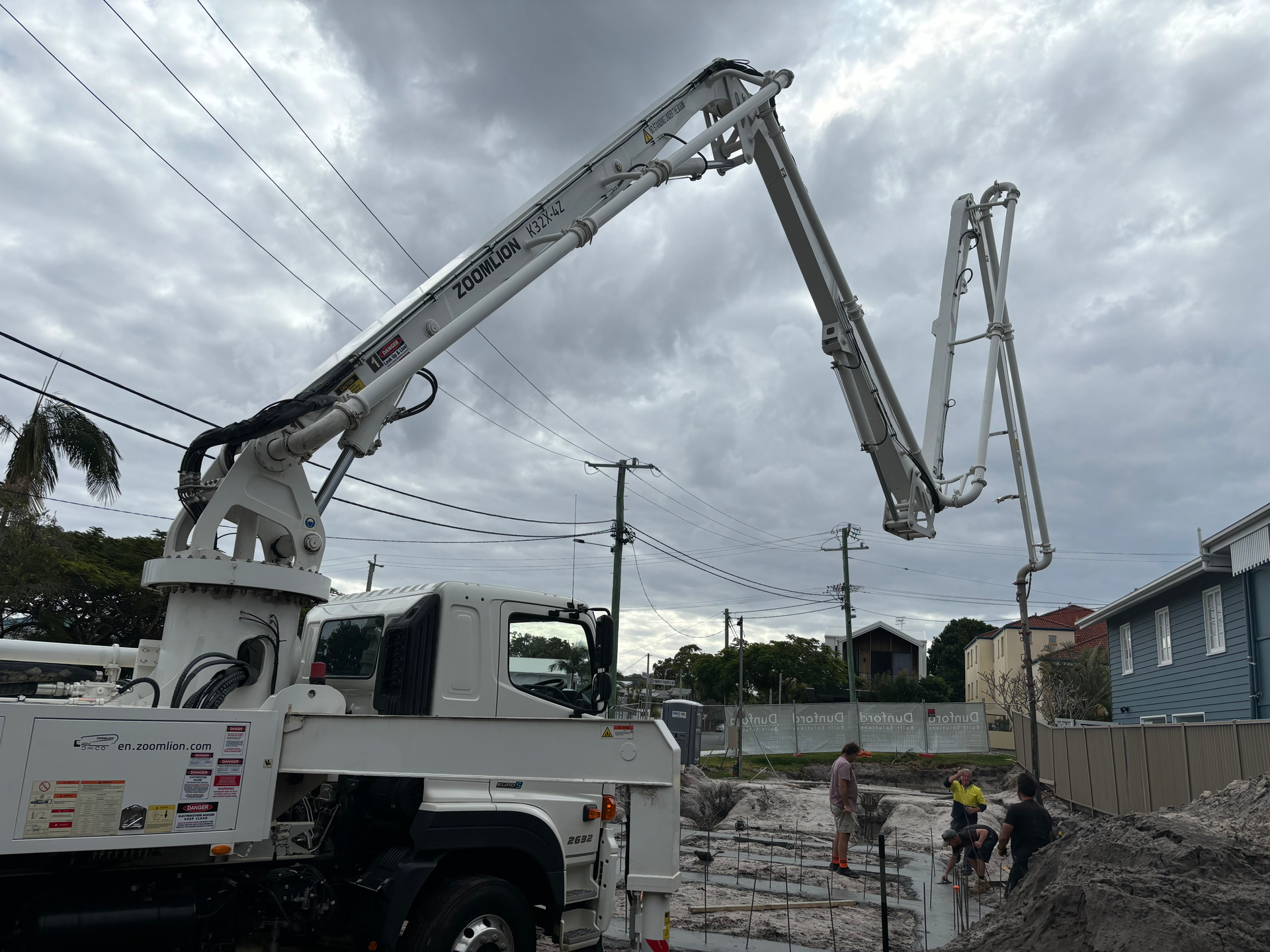 Concrete pump truck extending boom over a construction site. Workers pour concrete, cloudy sky.