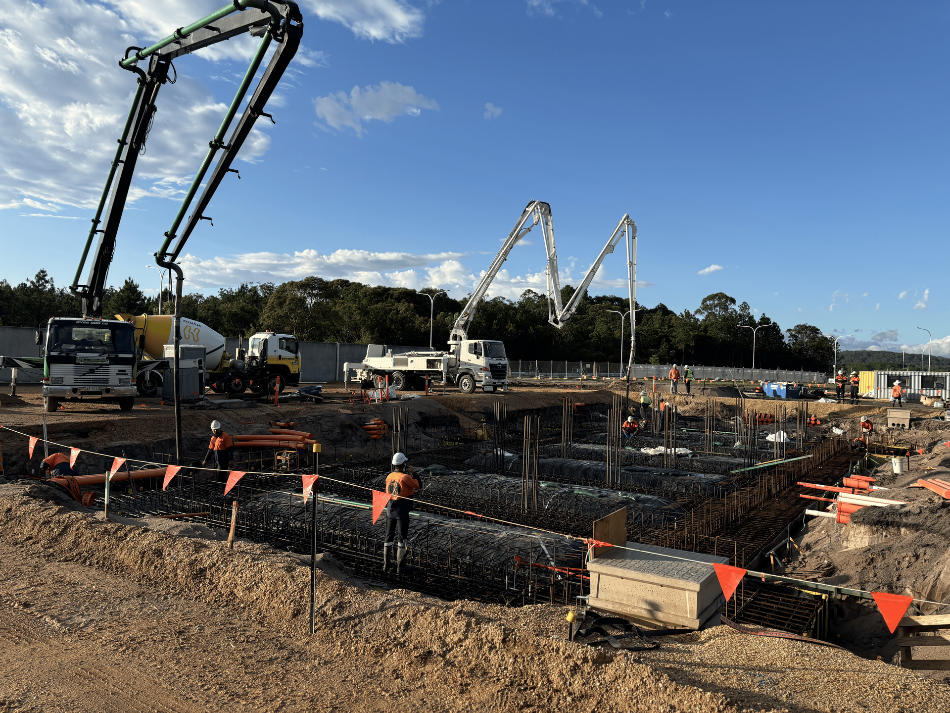 Construction site: concrete being poured into a foundation, using pump trucks; workers present.