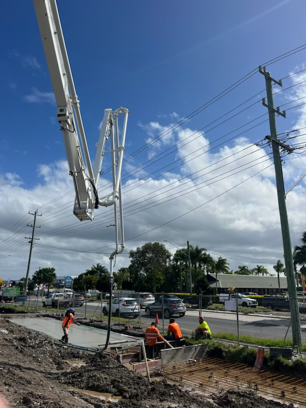 Concrete pump pouring concrete on road construction site; workers in orange vests nearby.