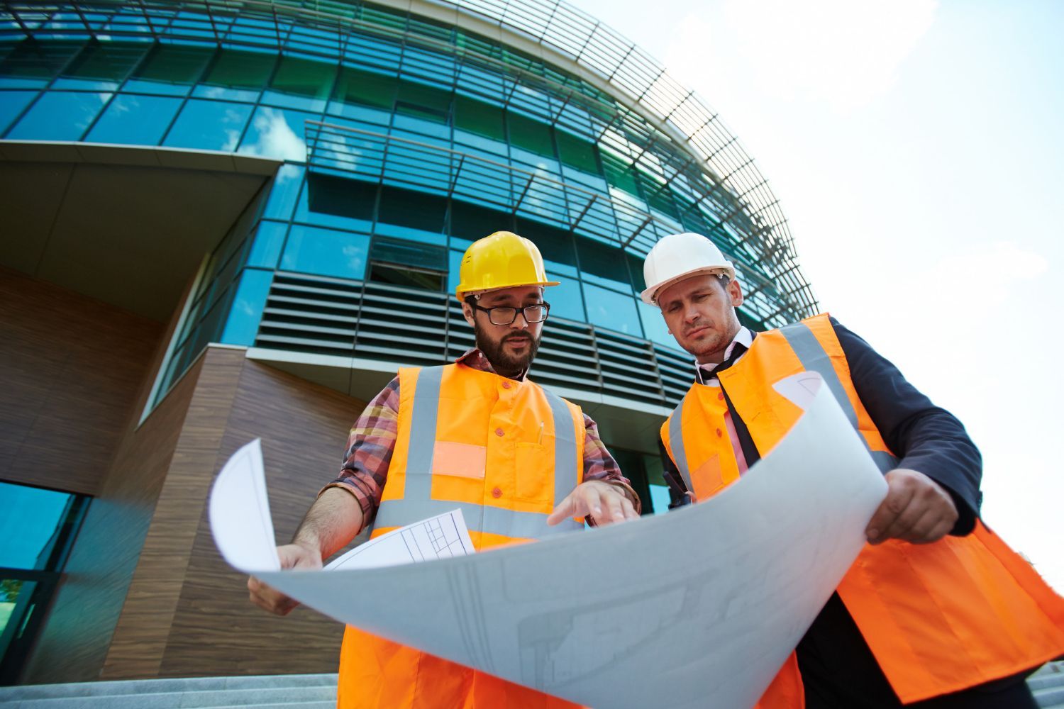 Two construction workers are looking at a blueprint in front of a building under construction.
