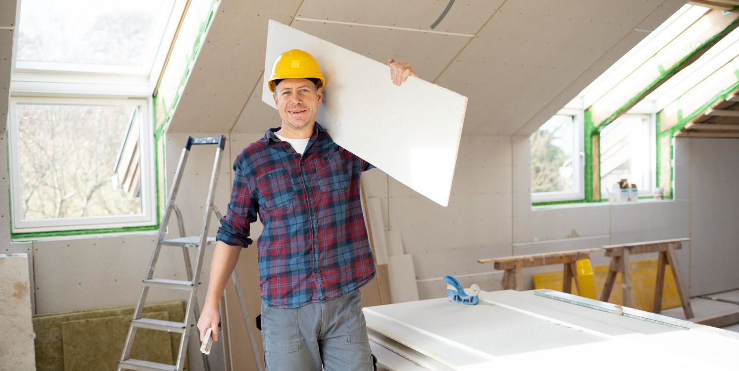 A man wearing a hard hat is carrying a piece of drywall on his shoulder.