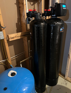Water filtration system with two black tanks, blue tank, and control panel, installed in a basement.