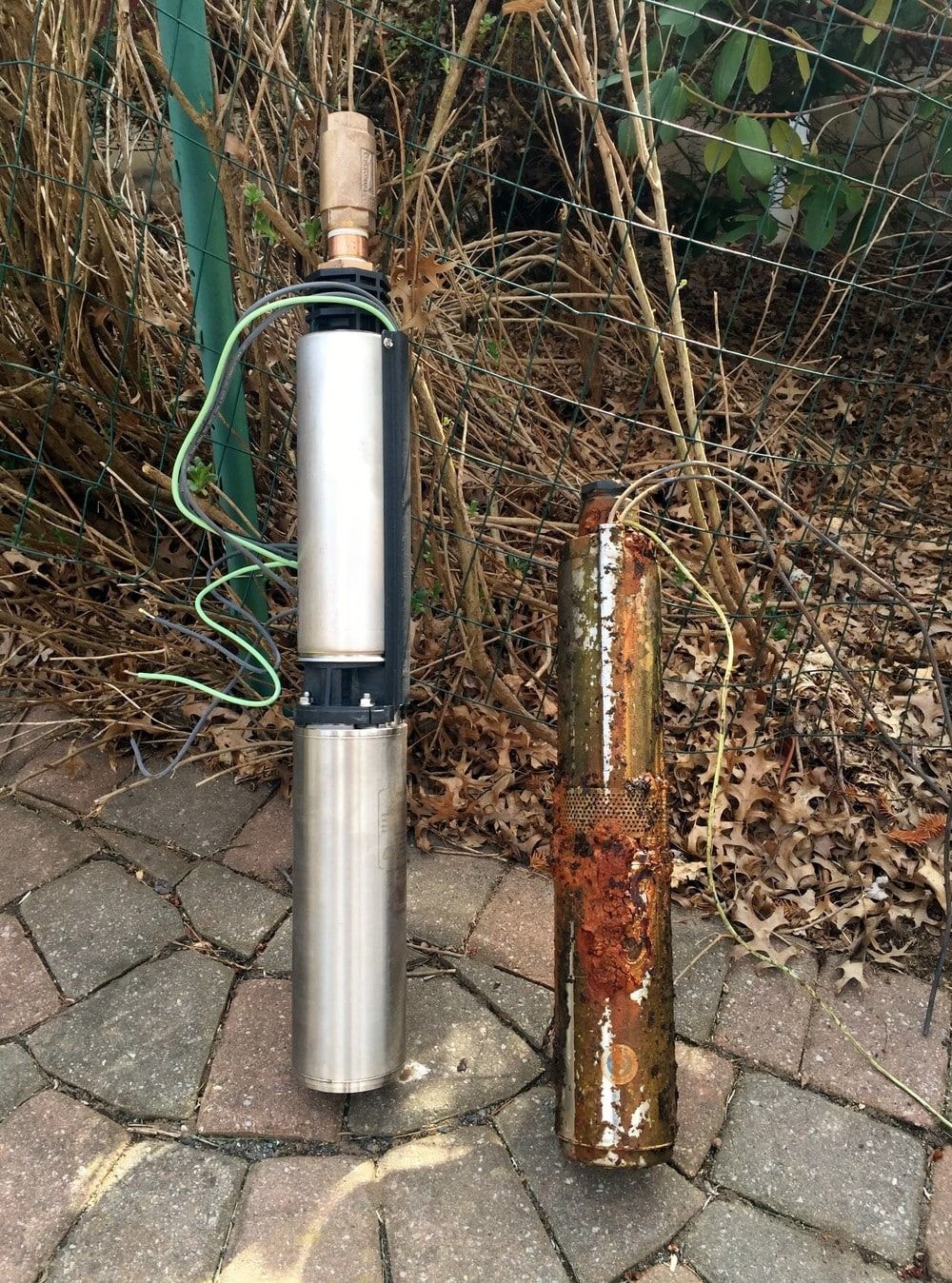 New and old well pumps side-by-side on a brick surface near brown foliage. The old one is rusty.