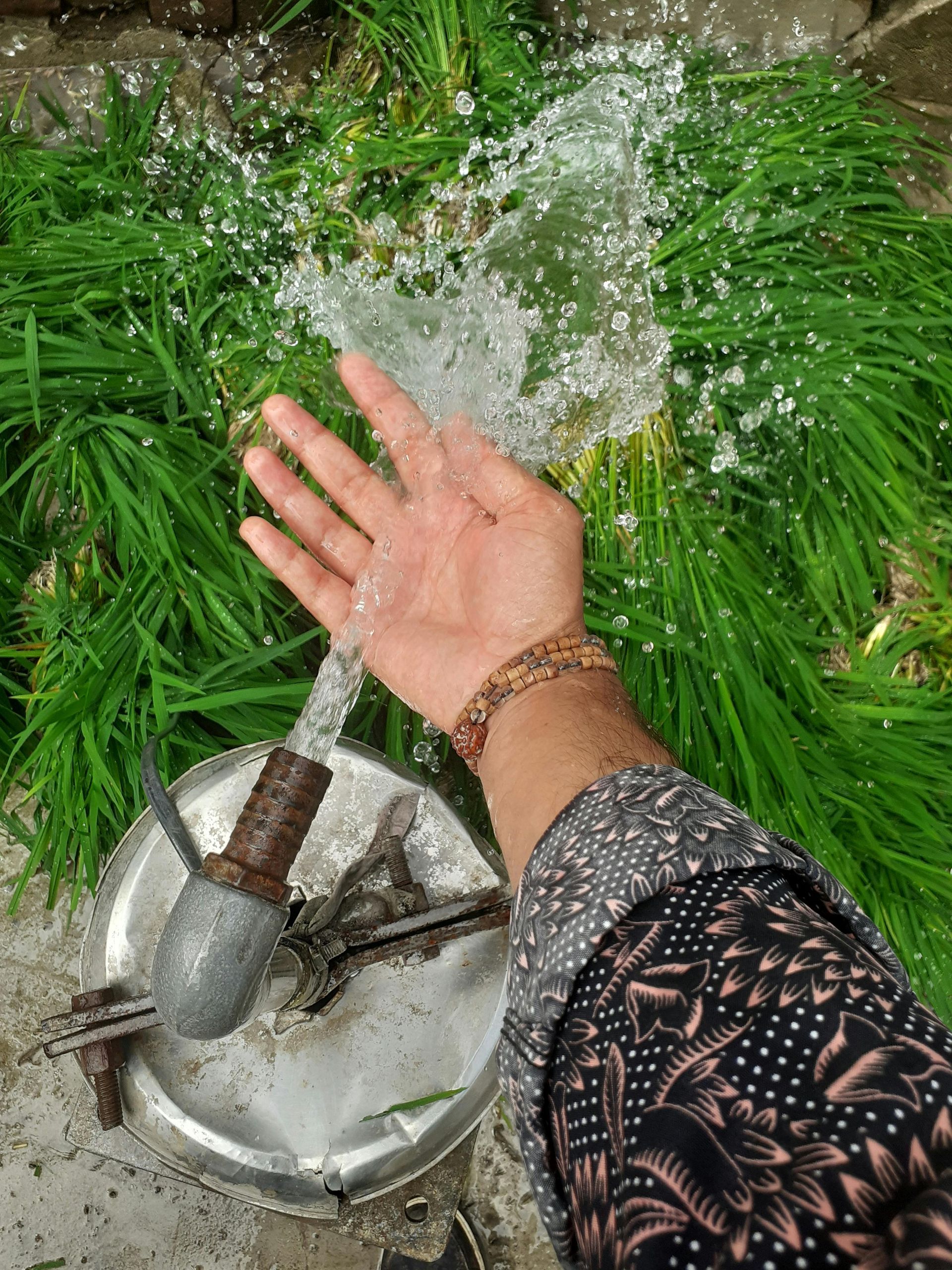 Hand under a stream of water, with grass background. Person wearing patterned sleeve and bracelets.