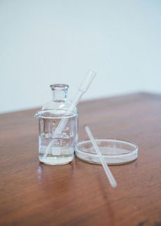 Clear glass bottle, pipette, and petri dish on a wooden table. Liquid inside bottle.