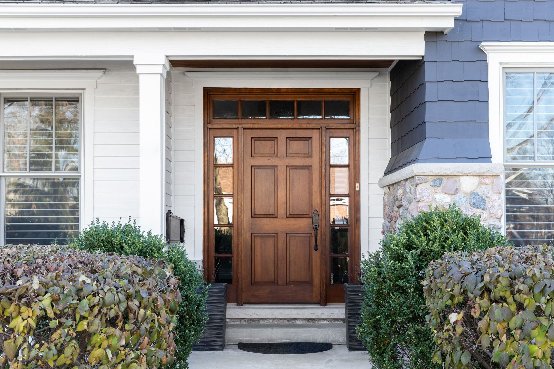 Wooden front entry door with glass sidelights, framed by white siding, stone accents, and shrubs.